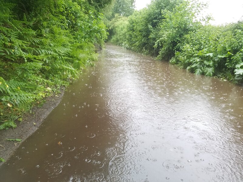 A large murky puddle with rain still falling heavily into it stretches across the road and for at least 40 metres ahead
